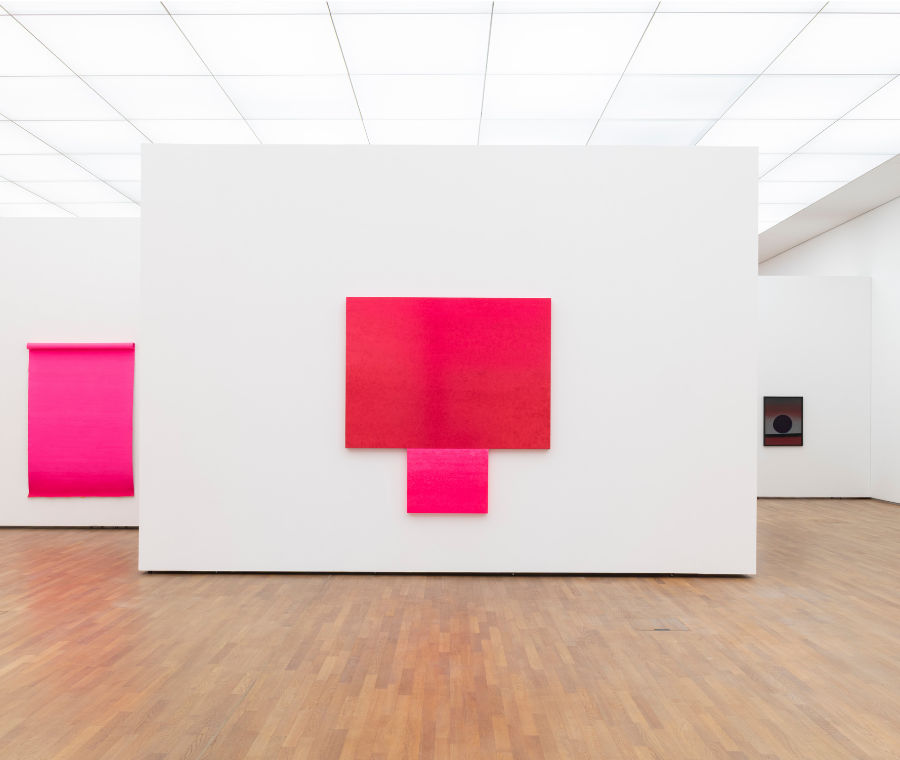 Ausstellungsansicht mit geometrischem rotem Kunstwerk von Rupprecht Geiger auf weißer Museumswand, umgeben von weiteren abstrakten Werken in hellem Ausstellungsraum